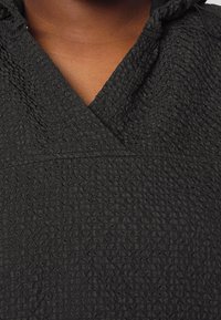 Zwarte hoodie met textuur, voorzien van een gekreukeld patroon, V-halsontwerp en een subtiele glans, met zichtbare stiksels nabij de halslijn.