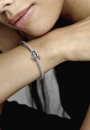 Silberarmband mit verdrehtem Design, ausgestattet mit einem herzförmigen Verschluss. Glatte Metalloberfläche, getragen an einem nackten Handgelenk vor einem neutralen Hintergrund.