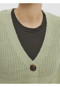 Jasnozielony dziergany kardigan z dekoltem w szpic, z zapięciem na jeden brązowy guzik. Nosi się go na ciemną, prążkowaną bluzkę. Gładka faktura.