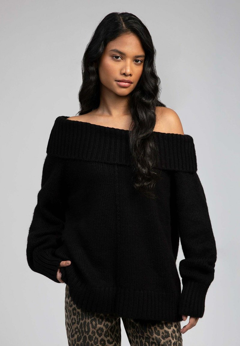 Pull en tricot noir à épaules dénudées avec col et poignets côtelés, affichant une coupe décontractée, porté avec un pantalon à imprimé léopard.