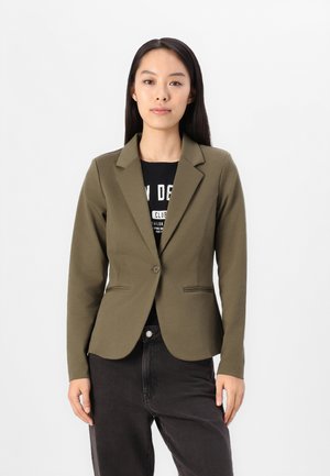 Olive green tailored blazer with notched lapels, zapięcie na jeden guzik oraz dwie przednie kieszenie, w połączeniu z czarną koszulką z grafiką i ciemnymi spodniami.