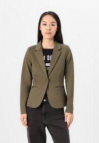 Olivgrön skräddarsydd blazer med slagkrage, enkel knappstängning och två framfickor, som bärs tillsammans med en svart grafisk t-shirt och mörka byxor.