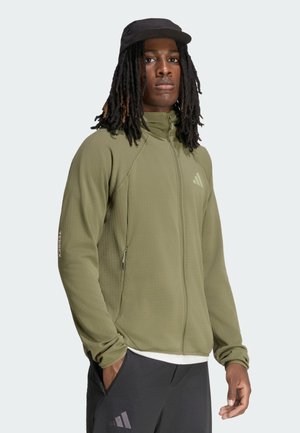 Mann trägt eine olivgrüne Jacke mit Reißverschluss und schwarze Mütze, mit Händen in den Taschen, steht vor einem schlichten hellen Hintergrund.