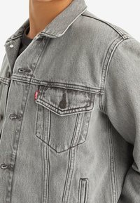 Graue Jeansjacke mit einem Kragen, einer Knopfleiste und zwei Brusttaschen mit einem roten Logoetikett. Der Stoff hat eine strukturierte Oberfläche.