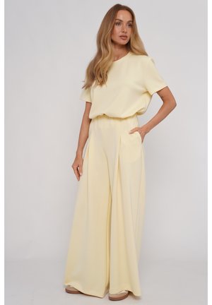 Kobieta z długimi blond włosami, ubrana w jasny żółty top z krótkim rękawem oraz spodnie z szerokimi nogawkami i wysokim stanem z kieszeniami, stojąca na jednolitym tle.