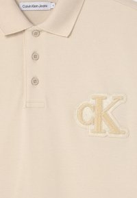 Beżowa koszulka polo z teksturowanej tkaniny, z trzema guzikami i dużym wyszywanym logo CK w jasnobeżowym kolorze po lewej stronie.
