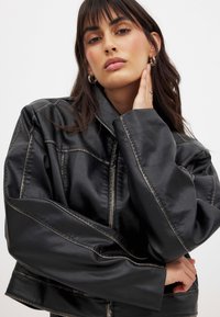 Jauna moteris su ilgais ruda plaukų, dėvinti juodą per didelį odinį švarką, sidabrines auskarus žiede ir žiedus, pozuojanti su ranka ant kaklo.