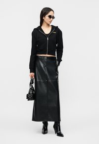 Γυναίκα με μαύρο κοντό φούτερ με φερμουάρ, μακριά μαύρη δερμάτινη φούστα, μαύρες μπότες με τακούνι, μικρή μαύρη τσάντα και γυαλιά ηλίου, στέκεται μπροστά σε λευκό φόντο.