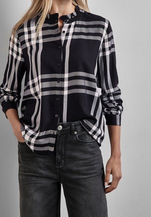 Kvinde iført sort-hvid ternet button-up skjorte med flæset krave og mørke jeans, hænderne i lommerne.