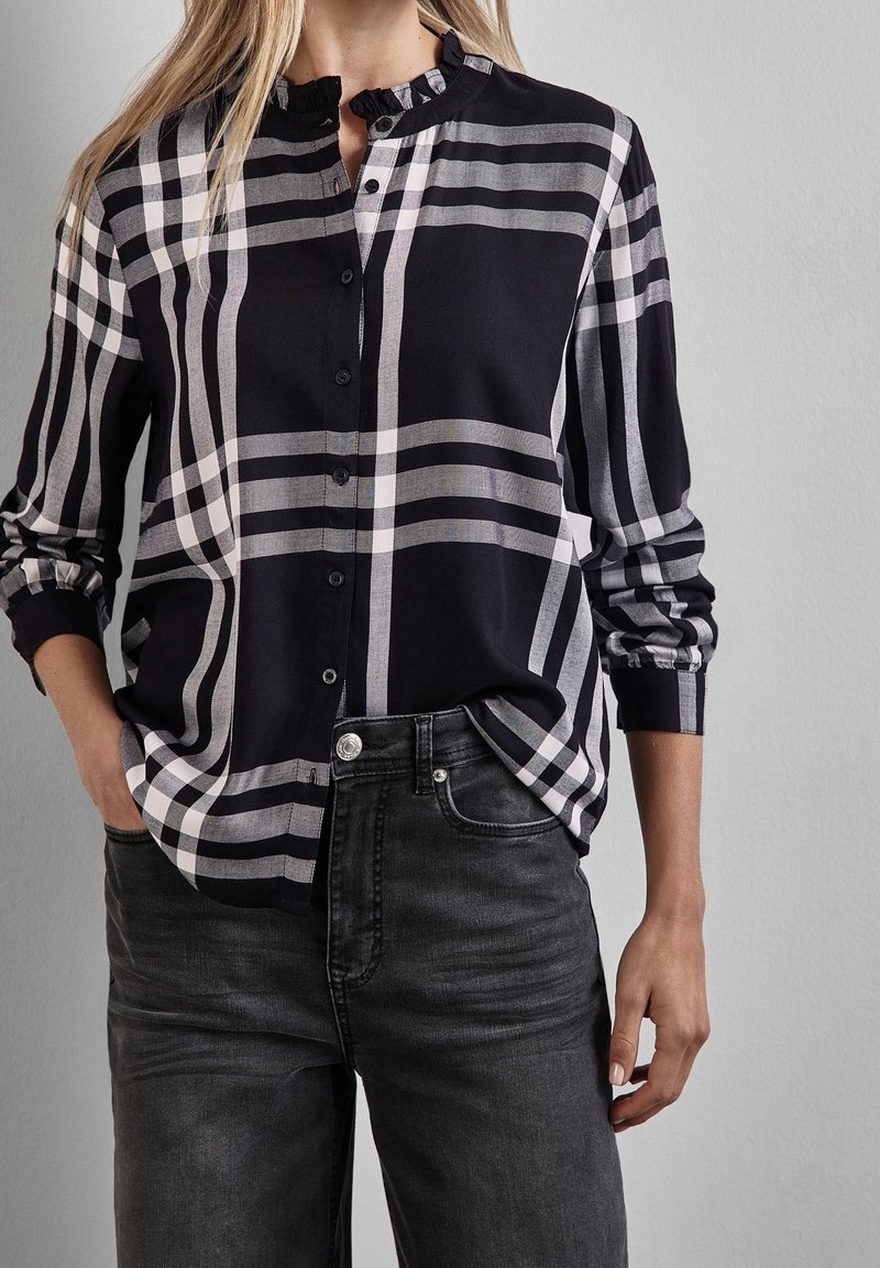 Femme portant une chemise à carreaux noir et blanc boutonnée avec un col à volants et un jean délavé foncé, mains dans les poches.