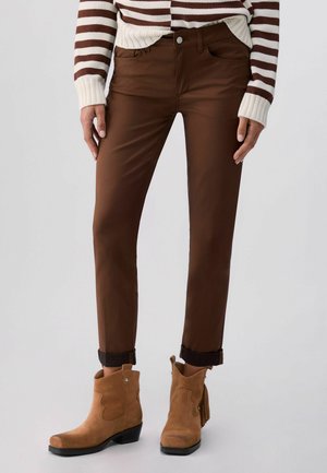 Persona che indossa pantaloni marroni arrotolati, stivaletti in camoscio color cuoio e un maglione a righe crema e marrone, in piedi contro uno sfondo chiaro.