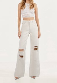 Světle šedé džíny s vysokým pasem a širšími nohavicemi s vybledlými detaily, které mají velké trhliny na kolenou a surové lemy, v kombinaci s bílým krajkovým crop topem.