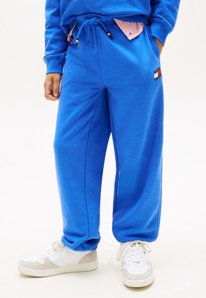 Persona vistiendo pantalones deportivos azul brillante con cordón y zapatillas blancas, de pie con las manos en los bolsillos sobre un fondo blanco.