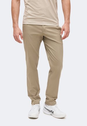 Άντρας που φοράει μπεζ casual παντελόνια και λευκά αθλητικά παπούτσια, στέκεται μπροστά σε ένα απλό φωτεινό φόντο.