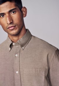 Lichtbruine, knoopsluiting shirt met een gestructureerde kraag, borstzak en een gladde textuur. Heeft vijf witte knopen aan de voorkant.