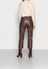 Pantalon en similicuir marron avec une coupe ajustée et une longueur jusqu'aux chevilles, assorti à des bottes à talons marron brillantes. Haut beige doux partiellement visible.
