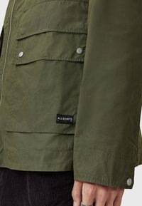 Veste vert olive en tissu lisse, dotée d'une fermeture éclair sur le devant, de poches à bouton-pression et d'une étiquette noire avec du texte blanc sur l'ourlet.