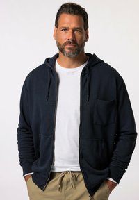 Navy zip-up hoodie with a drawstring, front pocket, and ribbed cuffs, layered over a white t-shirt and paired with beige pants.