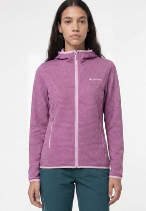 Jeune femme portant une veste à capuche violette zippée et un pantalon bleu sarcelle foncé, posant sur un fond blanc uni.