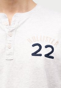 Φωτεινό γκρι πουκάμισο henley με τρία κουμπιά και κεντημένο κείμενο "HOLLISTER" σε μπεζ χρώμα πάνω από το ναυτικό μπλε νούμερο "22" στο στήθος.