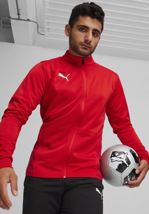 Rode zip-up jas met lange mouwen en wit Puma-logo. Heeft een gladde textuur. Zwarte broek met Puma-logo en een grijze voetbal.