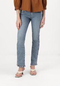 Vaqueros de mezclilla azul con corte ajustado y dobladillo ligeramente acampanado. Llevados con sandalias de tacón beige. La parte superior es una blusa marrón de corte holgado.