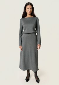 Vestito lungo grigio a maniche lunghe con scollo a barca, vita aderente e gonna fluida. Texture liscia senza fantasie, abbinato a tacchi neri.