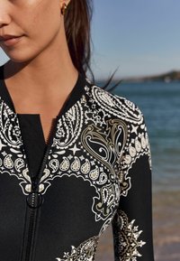 Čierny dlhý neopren s prepracovaným bielym a zlatým paisley vzorom, ktorý má predný zips a priliehavý strih.
