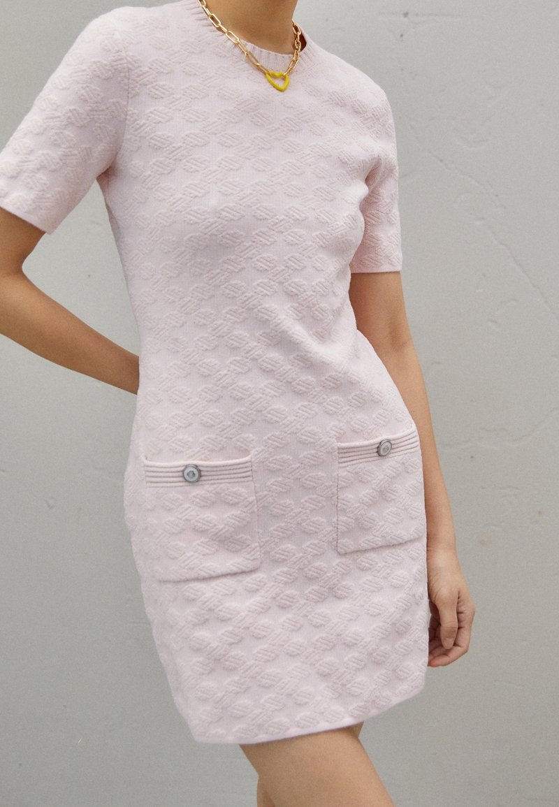 Femme portant une robe à manches courtes texturée rose clair avec des poches à boutons sur le devant et un collier chaîne doré avec un pendentif en forme de cœur jaune.