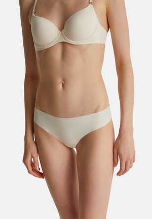 Femme portant un soutien-gorge moulé beige et une culotte bikini assortie sans coutures, sur un fond clair uni.