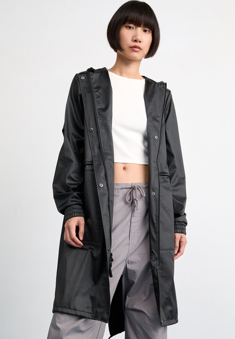 Black long coat in smooth, shiny fabric with a hood, elastic cuffs, and two front pockets, worn over a white crop top and gray pants.