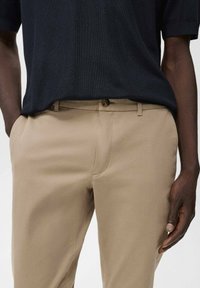 Beige broeken van een gladde stof, slim fit, met zijzakken. De taille heeft een metalen knoop en riemlussen, met daarboven een marineblauwe blouse.