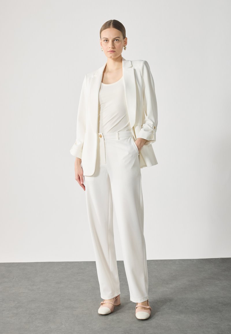 Completo bianco su misura con giacca a un bottone, pantaloni a gamba larga e top aderente, abbinato a scarpe con tacco block color crema.