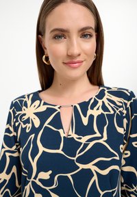 Navybluse mit floralem beigen Muster, Schlüssel-ausschnitt-Detail und glatter Stofftextur. Goldene Creolen passen perfekt zum Design.