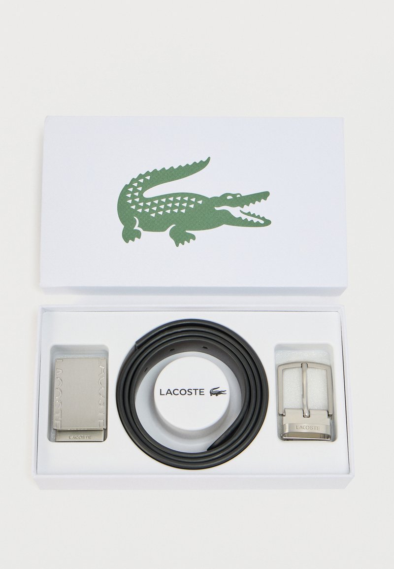 Ceinture noire avec boucle circulaire, ferrures en argent et plaque métallique marquée "LACOSTE", dans une boîte blanche ornée d'un logo crocodile vert.