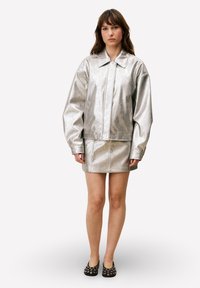 Veste métallisée argentée avec col, manches longues et taille cintrée, assortie à une jupe coordonnée. Chaussures plates noires avec des accents.