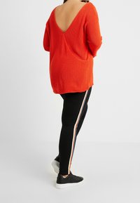 Maglioncino arancione brillante a maglia con design a V profonda sulla schiena, abbinato a leggings neri con strisce multicolori lungo i lati. Sneakers nere.