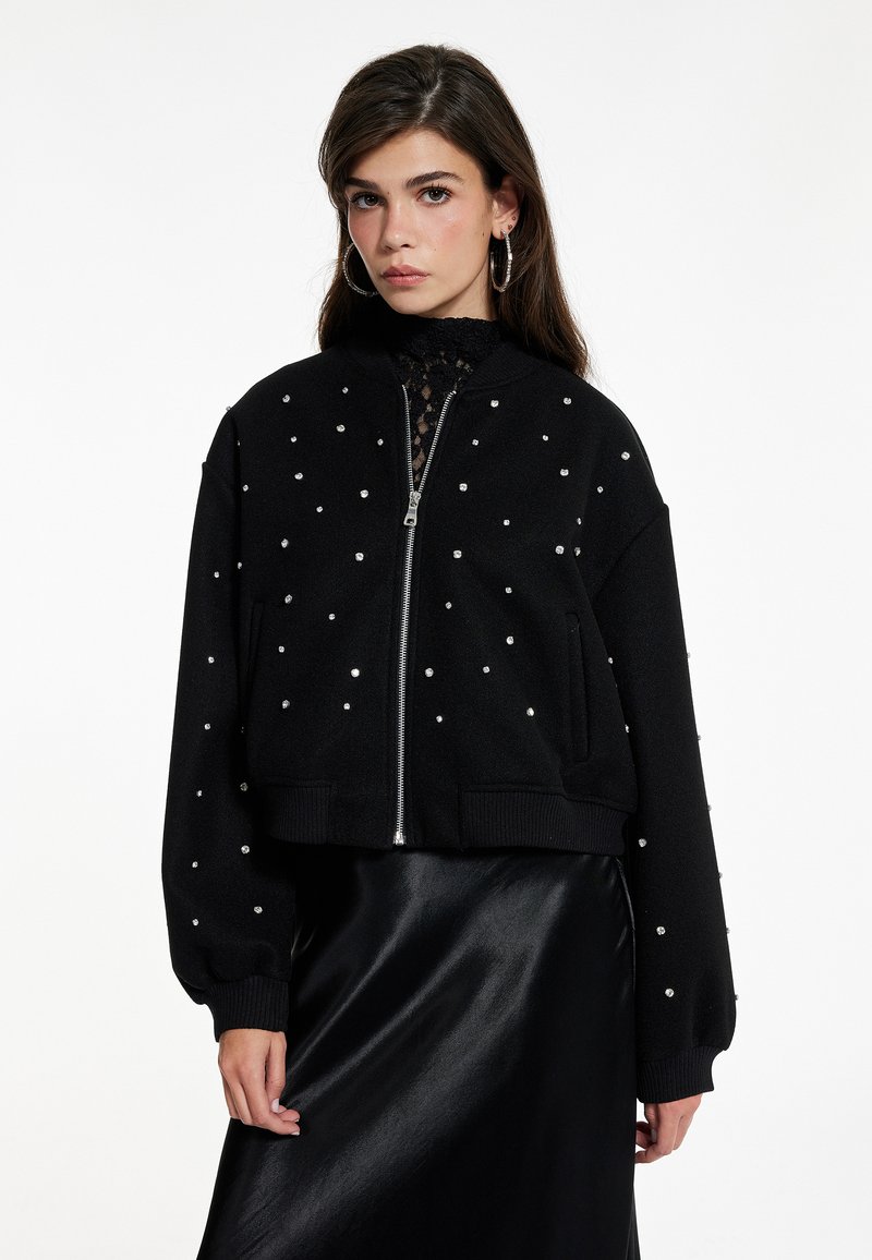 Schwarze Bomberjacke mit Reißverschluss und kleinen silbernen Nieten verziert. Lange Ärmel und gerippter Saum. Über einem schwarzen Satinrock getragen.