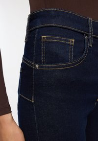 Granatowe dżinsy mają żółte stebnowanie, wysoki stan oraz kieszeń z detalem w postaci metalowych nitów. Tkanina ma lekko chropowatą strukturę.