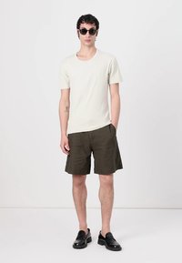 Light beige short-sleeve t-shirt, olive green shorts, and black shoes; model wearing black sunglasses, standing against a plain background.