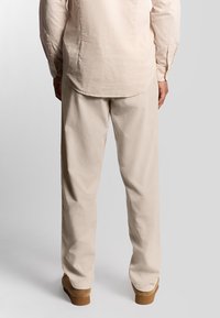 Mand iført beige bukser, beige langærmet skjorte og lysebrune sko, stående mod en ensfarvet lys grå baggrund.