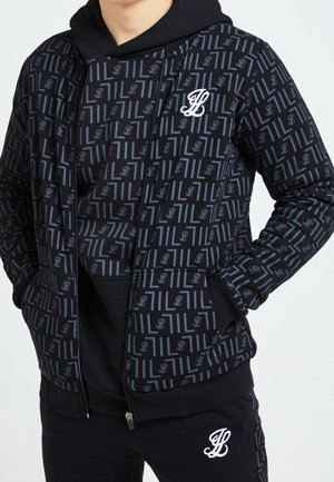 Mand iført sort hættetrøje og jakke med gråt geometrisk mønster og hvidt broderet kursivt "JL" logo på brystet og bukser.