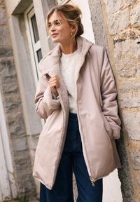 Giacca imbottita color beige chiaro con fodera in morbido pelliccia, colletto alto e chiusura con zip. Abbinata a un maglione di crema con texture e pantaloni in denim scuri.