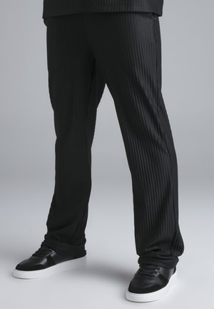 Personne portant un pantalon noir à fines rayures et des baskets noires avec des semelles blanches debout sur un sol gris clair.