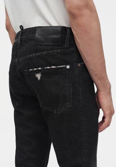 Homme portant un jean noir déchiré avec un patch en cuir du logo DSQUARED2 à la taille et une chemise blanche rentrée.