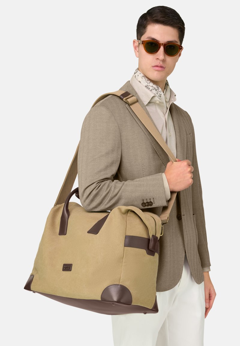 Homme portant un blazer beige, une chemise blanche, un foulard à motifs autour du cou, des lunettes de soleil, et portant un grand sac de voyage beige et marron avec bandoulière.