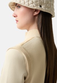 Beige gestrickter Pullover mit hohem Kragen, der gepolsterte Schulterakzente und eine glatte Textur aufweist. Kombiniert mit einem passenden gepolsterten Bucket Hat.