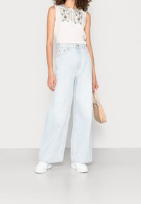 Jean large bleu clair à taille haute, associé à un haut sans manches blanc avec broderie florale et un col à nouer.