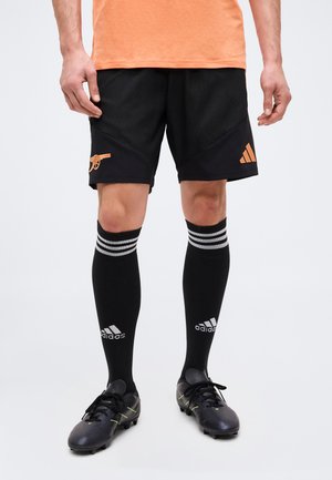 Muškarac obučen u crne Adidas nogometne kratke hlače s narančastim logotipima, crne Adidas čarape do koljena i crne kopačke stoji na bijeloj pozadini.