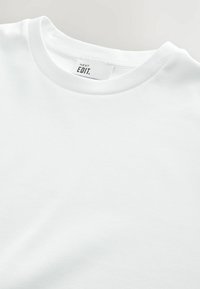 Weißes Baumwoll-T-Shirt mit geripptem Rundhalsausschnitt und normalem Schnitt. Das Etikett trägt die Aufschrift "NEXT EDIT." Der Stoff wirkt glatt und unstrukturiert.